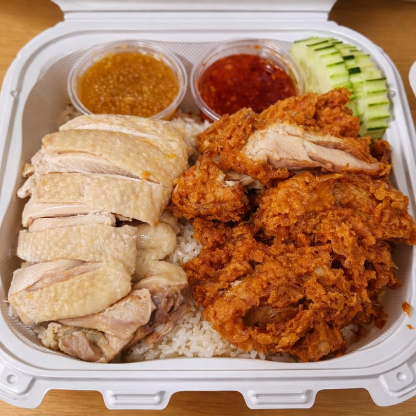 Hainanese chicken rice with steamed and fried chicken, two sauces, and cucumber slices in a takeout container.