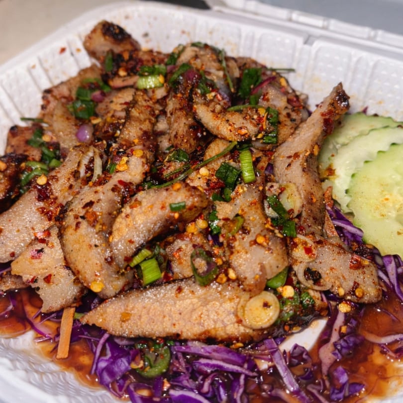 Spicy grilled pork salad with red cabbage, green onions, and cucumber slices in a white takeout container.