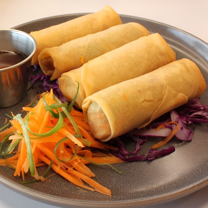 Four golden spring rolls with shredded carrots, red cabbage, and a dipping sauce on a gray plate.